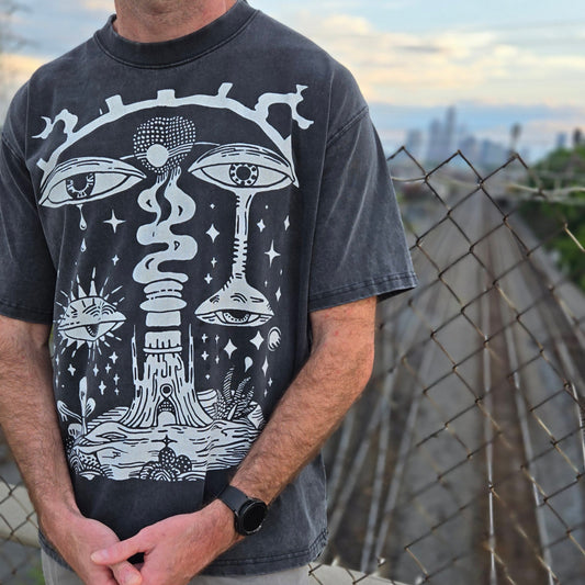 Male model wearing charcoal wash Paradoxical Perspective tee front view cropped at waist with city skyline behind fence