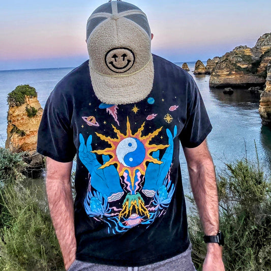 Man in smiley face cap wearing black Yin Yang cosmic graphic tee at coastal cliff overlook