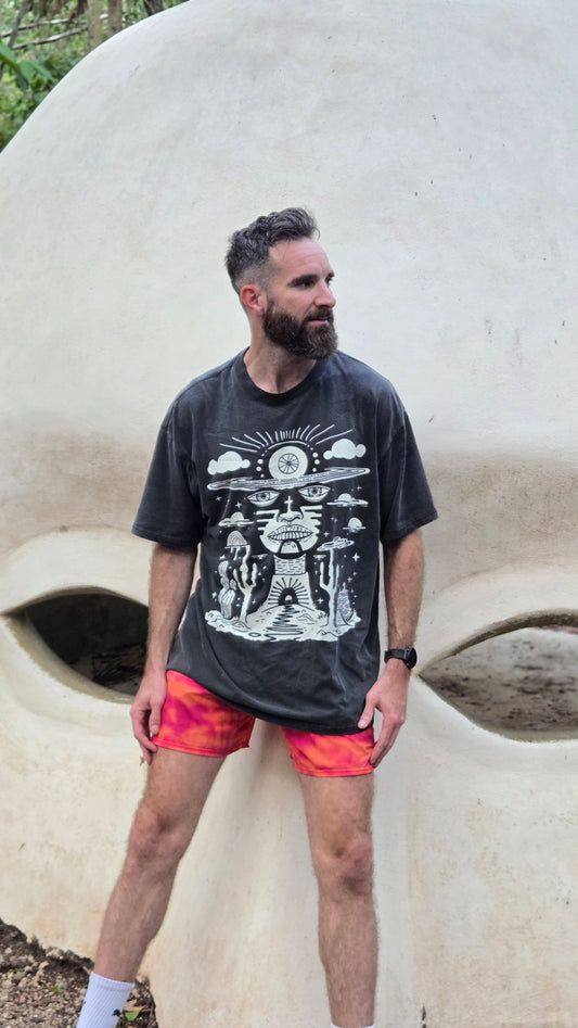 Bearded man wearing Deserted Visions tee with orange shorts standing against large stone face sculpture