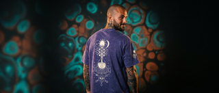 Back of the violet Celestial Synthesis snow wash heavyweight tee with a tree of life print, in front of a liquid light projection