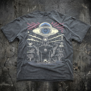 Faded charcoal Extraterrestrial fluffy vintage heavyweight tee on cracked concrete, printed in cream, pink and blue with a ringed planet, meditating figure and a mystical desert scene.