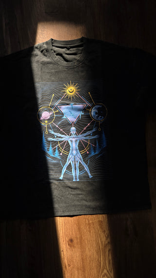 A black graphic t-shirt laid flat on a wooden floor, featuring a detailed celestial design with a sun, moon, and human figure in blue, white, and yellow.