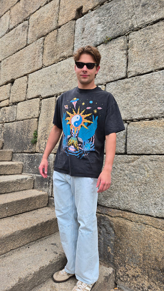 Man in sunglasses wearing black Yin Yang graphic tee with light jeans leaning against stone wall