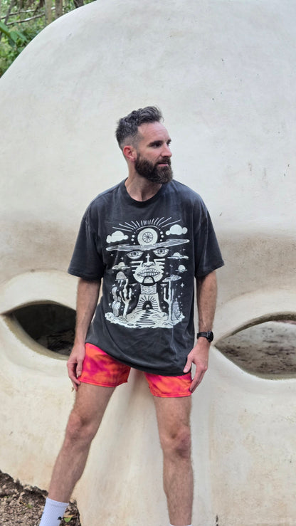Bearded man wearing Deserted Visions tee with orange shorts standing against large stone face sculpture