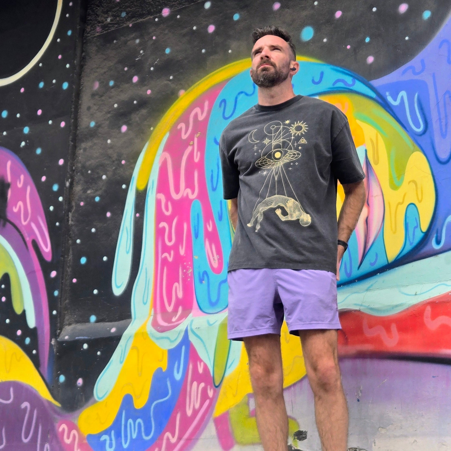 Man wearing Dimensional Drift charcoal tee with gold cosmic design standing against colorful mural