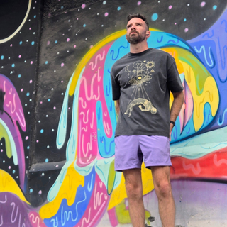 Man wearing Dimensional Drift charcoal tee with gold cosmic design standing against colorful mural