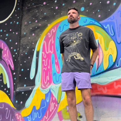 Man wearing Dimensional Drift charcoal tee with gold cosmic design standing against colorful mural