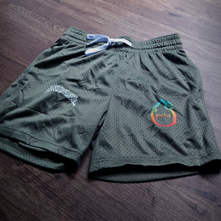 Dark olive green mesh athletic shorts with a white drawstring, laid flat on a dark wood floor. The shorts feature a white graphic on the left leg and a colorful circular logo on the right leg.