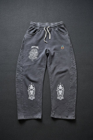 A pair of dark gray sweatpants with white graphic prints on the legs and a white drawstring, laid flat on a light wood floor.