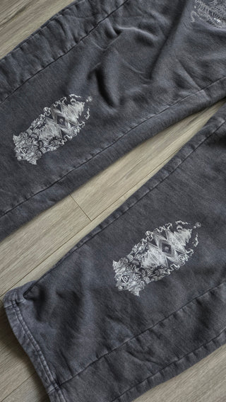 Washed gray Spirit Molecule barrel leg sweatpants laid on a light wood floor, showing the mirrored totem prints running down both shins