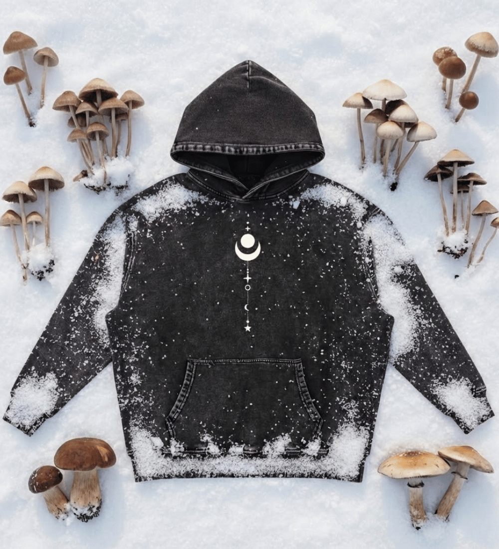 Black Existential snow wash hoodie with moon graphic laid flat on snow with mushrooms