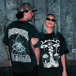 Two people standing in front of a weathered metal wall, each wearing a oversized graphic T-shirt. The person on the left shows the back of the shirt with white gothic text and an eye design, while the person on the right shows the front of the shirt with a white psychedelic mushroom and eye graphic.