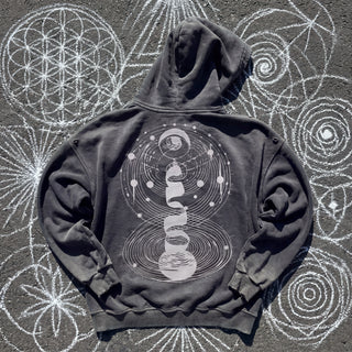 Cosmic Connection ultra heavyweight zip hoodie in washed Black, back view, laid on dark asphalt covered in white chalk sacred geometry, showing the white celestial artwork of a crescent moon, orbital rings and stacked spirals down the back.