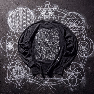 Back of the Embrace The Chaos acid wash oversized long sleeve tee in Black Beauty, styled on dark asphalt covered in white chalk sacred geometry, showing a white line-art panel of intertwined serpents.