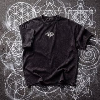 Front of the Embrace The Chaos Patakota collab tee in black snow wash on chalk sacred geometry, with a small white hypnotic eye and coiled serpent graphic at the chest.