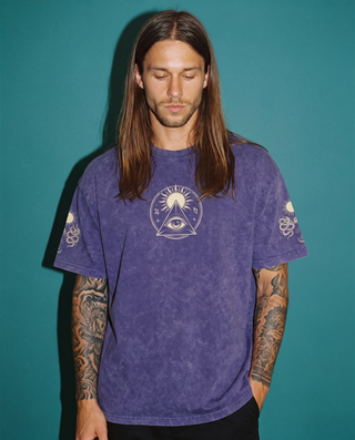 Model wearing the Celestial Synthesis snow wash oversized heavyweight tee in violet purple against a teal backdrop, printed on the chest with a cream all-seeing eye inside a triangle beneath a radiating sun, with snake and sun motifs on both sleeves.
