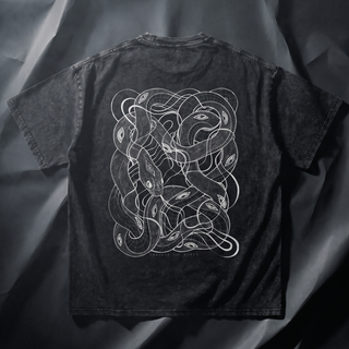 Back view of the Embrace The Chaos Patakota collab snow wash tee in black, laid on grey fabric and printed with a large white line-art tangle of intertwined serpents and eyes.