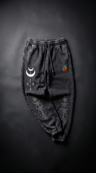 Charcoal snow wash Existential heavyweight joggers laid flat on dark stone, with an elasticated drawstring waist and cuffed ankles, printed with a white crescent moon and hanging stars on one thigh and sacred geometry line art down both lower legs.