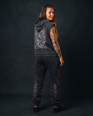 Back view of a model 5'4" and 125 lb wearing the charcoal Embrace The Chaos snow washed boxy hooded tank top in size M, hood down, showing the dropped sleeveless armholes and the white tangled serpent line art printed across the back, styled with charcoal Existential snow wash joggers with cuffed ankles.