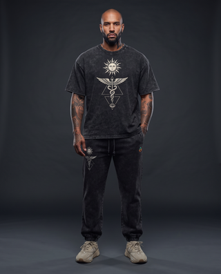 Model 5'9" and 185 lb wearing the full Inner Alchemy Snow Wash Set in black — charcoal snow wash oversized tee in size XL with a cream sun face, winged caduceus and all-seeing eye centred on the chest, over the matching 420 GSM black snow wash heavyweight joggers with the sun and caduceus leg print.