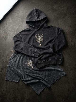 A dark washed black hooded sweatshirt and matching shorts set laid flat on a dark textured surface. Both pieces feature a small gold-toned celestial eye graphic on the front and thigh.