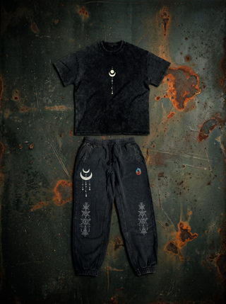 Existential Snow Wash Set laid flat on dark weathered steel — black snow wash 250 GSM oversized heavyweight tee with a cream crescent moon and celestial stack chest print, above the matching 420 GSM black snow wash heavyweight joggers with crescent moon and sacred geometry leg prints.