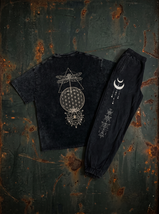 Existential Snow Wash Set, back view on dark weathered steel — black snow wash heavyweight tee printed in cream with a dragonfly over a Flower of Life sacred geometry mandala, beside the matching black snow wash joggers with crescent moon and alchemical line art down the leg.