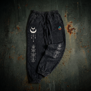 Existential 420 GSM black snow wash heavyweight joggers laid flat on dark weathered steel, showing the drawstring elastic waistband, side pockets, ribbed ankle cuffs and the cream crescent moon and sacred geometry prints down the leg.