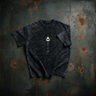 Existential black snow wash oversized heavyweight tee in a styled flat lay on dark weathered steel, one sleeve folded across the body, showing the mottled mineral snow wash and the small cream crescent moon and celestial stack chest print.