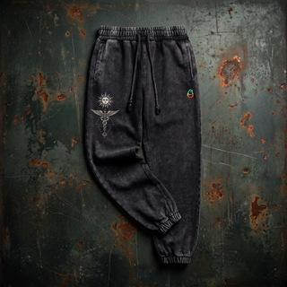 Inner Alchemy 420 GSM charcoal snow wash heavyweight joggers laid flat on dark weathered steel, showing the drawstring elastic waistband, side pockets, ribbed ankle cuffs and the cream sun face and winged caduceus print on the thigh.