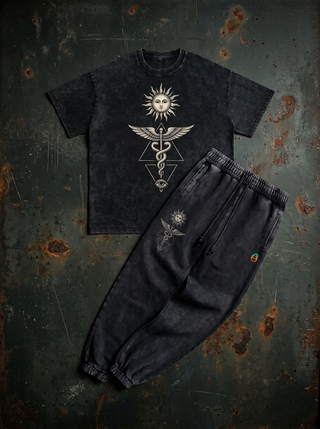 Inner Alchemy Snow Wash Set laid flat on dark weathered steel — charcoal snow wash oversized heavyweight tee with a cream radiant sun face, winged caduceus and all-seeing eye chest print, above the matching 420 GSM snow wash heavyweight joggers with the sun and caduceus leg print.
