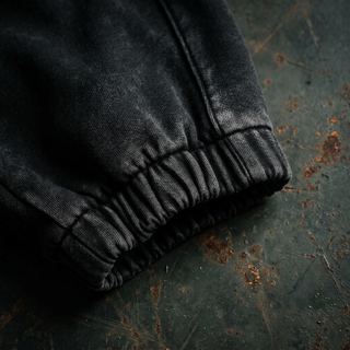 Close-up of the ribbed elasticated ankle cuff on the black snow wash Existential heavyweight joggers, shot on rusted dark steel.