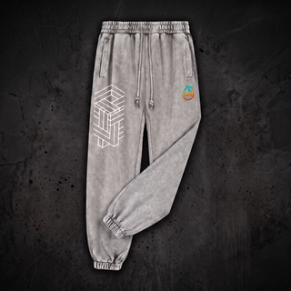Paradigm 420 GSM heavyweight joggers in cool Light Gray mineral snow wash, laid on near-black marble with one leg folded across the other at the knee, showing the drawstring elastic waistband, slanted side pockets, ribbed ankle cuffs, the white impossible-geometry cube printed on the thigh and the pH flame logo on the hip.