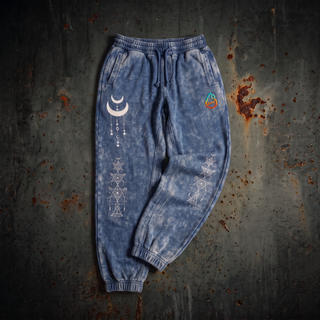 Existential 420 GSM heavyweight joggers in Royal Blue mineral snow wash, laid flat on dark weathered steel, showing the drawstring elastic waistband, slanted side pockets, ribbed ankle cuffs, the cream crescent moon and sacred geometry prints down the leg and the pH flame logo on the hip.