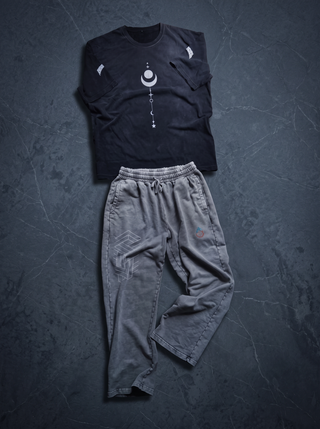 The Existential x Paradigm Set laid out on dark charcoal slate: the 260 GSM hand-frayed vintage wash tee in faded black above the 420 GSM snow washed baggy sweatpants in Light Gray, showing the impossible-geometry leg print and the pH flame logo.