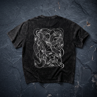 Back of the Embrace The Chaos Patakota collab tee in black snow wash, crumpled into a styled flat lay on dark charcoal slate, printed in white line art with a dense tangle of intertwined serpents and eyes.