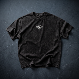 Front of the Embrace The Chaos Patakota collab tee in black snow wash, crumpled into a styled flat lay on dark charcoal slate, with the small white eye-and-saucer graphic printed at the chest and the boxy drop-shoulder cut visible.