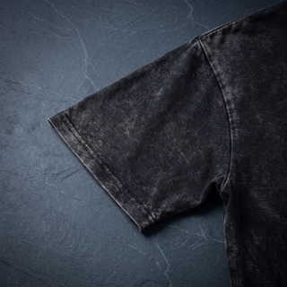 Close-up of the short sleeve hem and shoulder seam on the 250 GSM black snow wash heavyweight tee, shot on dark charcoal slate.