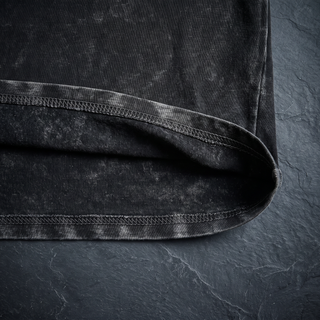 Close-up of the doubled bottom hem and overlock stitching on the 250 GSM black snow wash heavyweight tee, shot on dark charcoal slate.