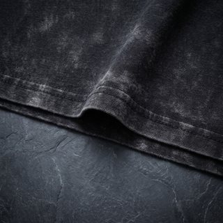 Close-up of the hem fold and topstitching on the 250 GSM black snow wash heavyweight tee, shot on dark charcoal slate.