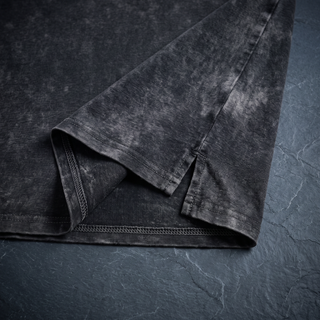 Close-up of the side seam, hem vent and chain stitching on the 250 GSM black snow wash heavyweight tee, shot on dark charcoal slate.