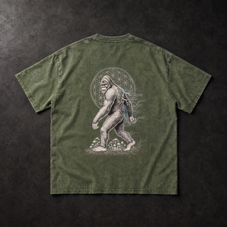 Studio flat lay of the Bean Green PsySquatch snow wash tee on charcoal concrete, back view, soft directional light across the moss green mineral wash, showing the full white sasquatch line art print with its Flower of Life mandala, teal mycelium and mushroom cluster