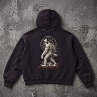 Studio flat lay of the black PsySquatch vintage wash boxy fleece hoodie on weathered charcoal concrete, back view, hood laid flat, showing the full white sasquatch line art print with its Flower of Life mandala, teal mycelium and mushrooms across the 390 GSM washed fleece