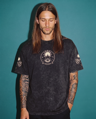 Model wearing the Celestial Synthesis snow wash oversized heavyweight tee in Black against a teal backdrop, printed on the chest with a cream all-seeing eye inside a triangle beneath a radiating sun, with snake and sun motifs on both sleeves.