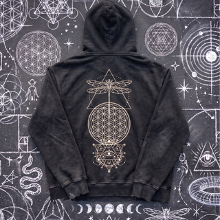 Black Existential snow wash hand-frayed heavyweight hoodie laid flat on wet chalk-covered pavement, showing the cream dragonfly, flower of life and all-seeing-eye celestial print centered on the back.