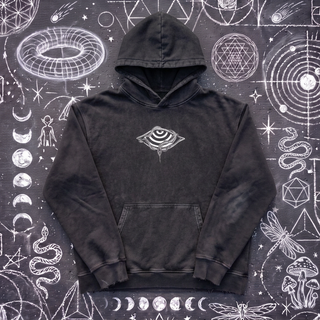 Black Beauty Embrace The Chaos snow washed loose cotton hoodie laid flat on wet chalk-covered pavement, front view with the small white striped eye in a wide diamond and a snake printed center chest, a Patakota collab.