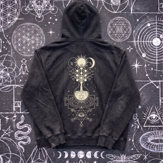 Black Celestial Synthesis snow wash hand-frayed heavyweight hoodie laid flat on wet chalk-covered pavement, showing the cream alchemical tree of life, flask and all-seeing eye print on the back.