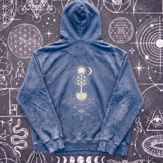 Royal blue Celestial Synthesis snow wash hand-frayed heavyweight hoodie laid flat on wet chalk-covered pavement, showing the cream alchemical tree of life, flask and all-seeing eye print on the back.