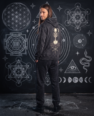 Model 6'0" and 165 lb wearing the black Celestial Synthesis snow wash hand-frayed heavyweight hoodie in size L, looking back over his shoulder in front of a chalkboard wall of white chalk sacred geometry, showing the cream alchemical tree of life print on the back, styled with black Existential snow wash joggers.