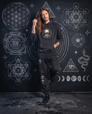 Model in the black Celestial Synthesis hoodie and joggers from the 3-Piece Set, leaning on a chalk sacred geometry wall.